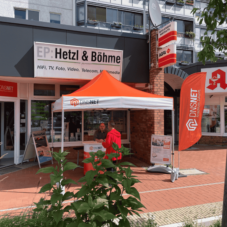 Ein DNS:NET-Infostand vor dem Elektronikgeschäft „EP: Hetzl & Böhme“ in einer urbanen Straße in Berlin-Brandenburg. Der Stand besteht aus einem orange-weißen Pavillon mit DNS:NET-Logo, einem Promotion-Tisch und einer orangen Beachflag. Zwei Männer unterhalten sich am Stand, einer trägt eine rote Jacke. Im Hintergrund sind Werbeschilder für Telekommunikation und Fotostudio sichtbar. Das Setting wirkt einladend und informiert über Glasfaser- und Telekommunikationsangebote für die Region. Im Vordergrund sind grüne Pflanzen zu sehen.