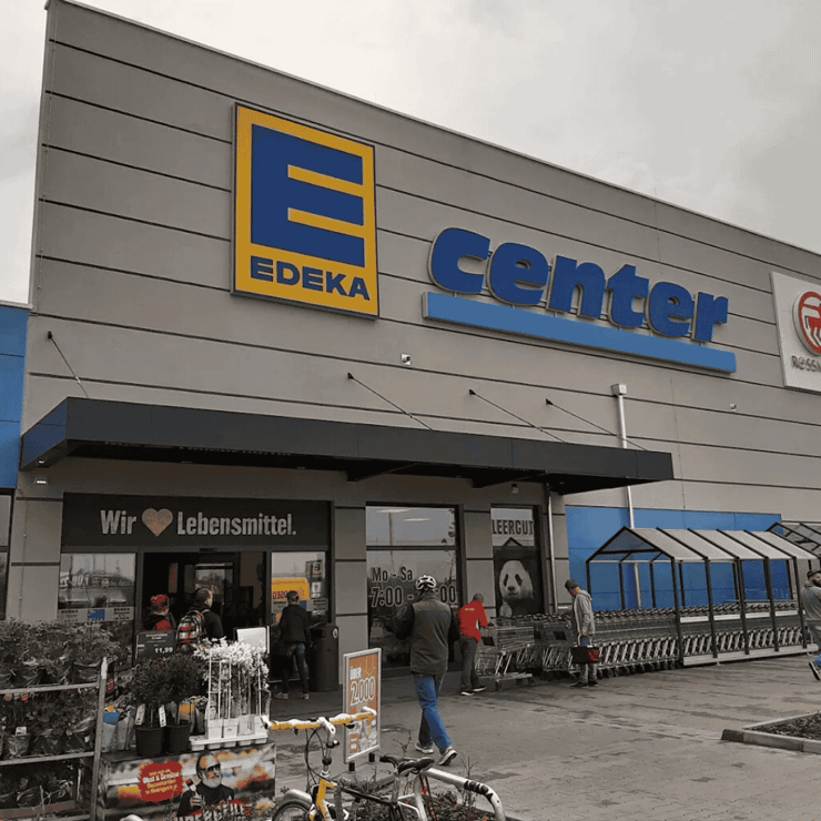 Fassade eines modernen EDEKA Centers mit großem gelbem EDEKA-Logo und blauem „Center“-Schriftzug, daneben ein ROSSMANN-Logo. Vor dem Eingang stehen Kunden, einige holen oder bringen Einkaufswagen zurück. Links befinden sich Pflanzen- und Blumenregale, rechts sind überdachte Wagenboxen für Einkaufswagen. Über dem Eingang steht der Slogan „Wir lieben Lebensmittel.“ Das Wetter ist bewölkt, die Atmosphäre wirkt geschäftig und einladend.