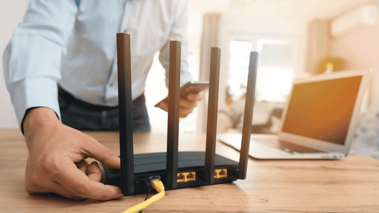 Mann verbindet WLAN-Router mit gelbem Ethernet-Kabel, Smartphone in der Hand, Laptop im Hintergrund.